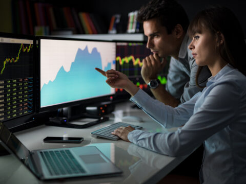 Two traders analyze market charts on multiple monitors, one pointing at a blue graph while the other watches closely.