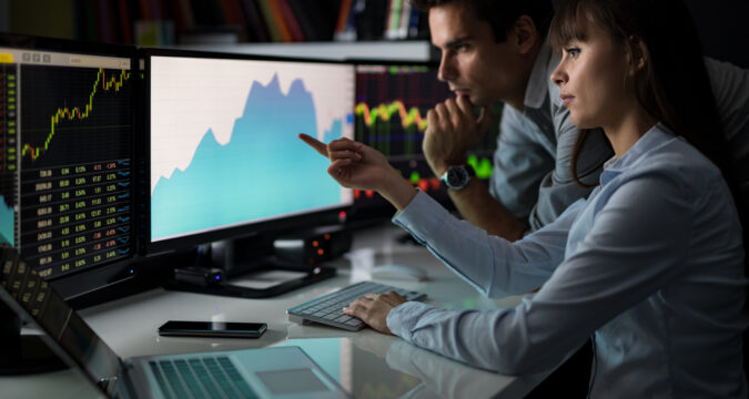 Two traders analyze market charts on multiple monitors, one pointing at a blue graph while the other watches closely.